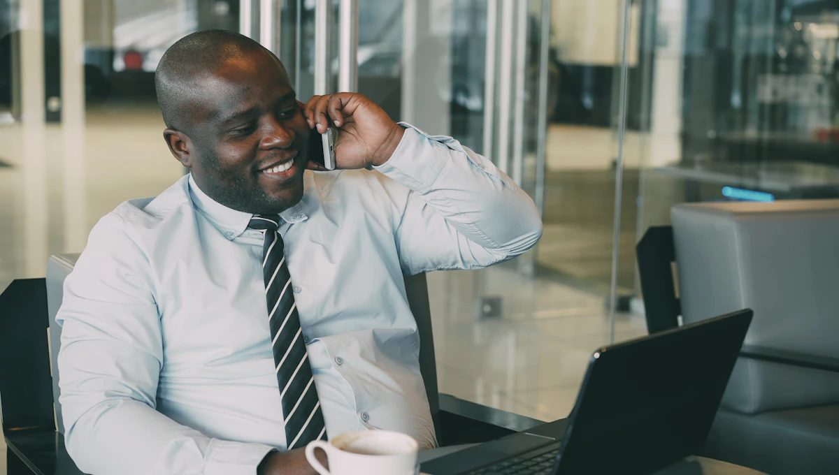 A smiling man in a shirt and tie on a phone call at a table, a laptop and a cup of coffee in front of him.