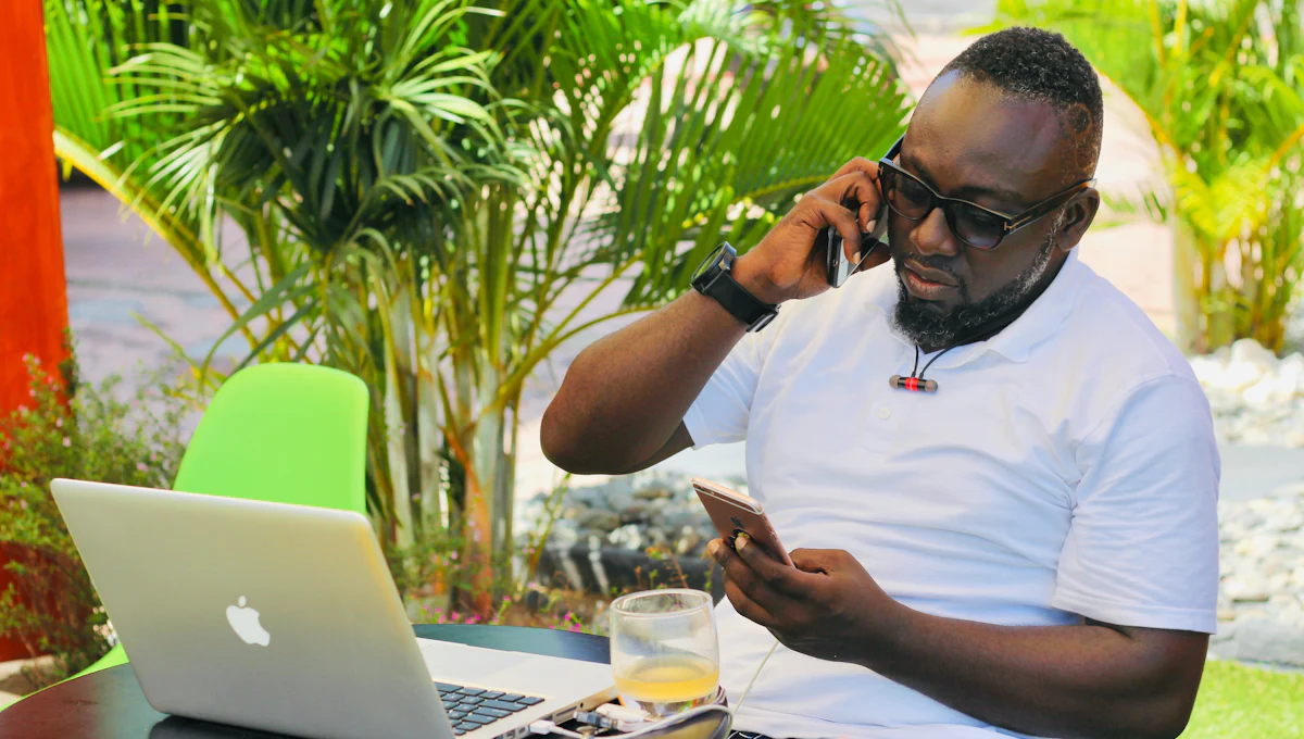 A business owner on a call outdoors, a laptop open on the table in front of him and a second phone in his hand.