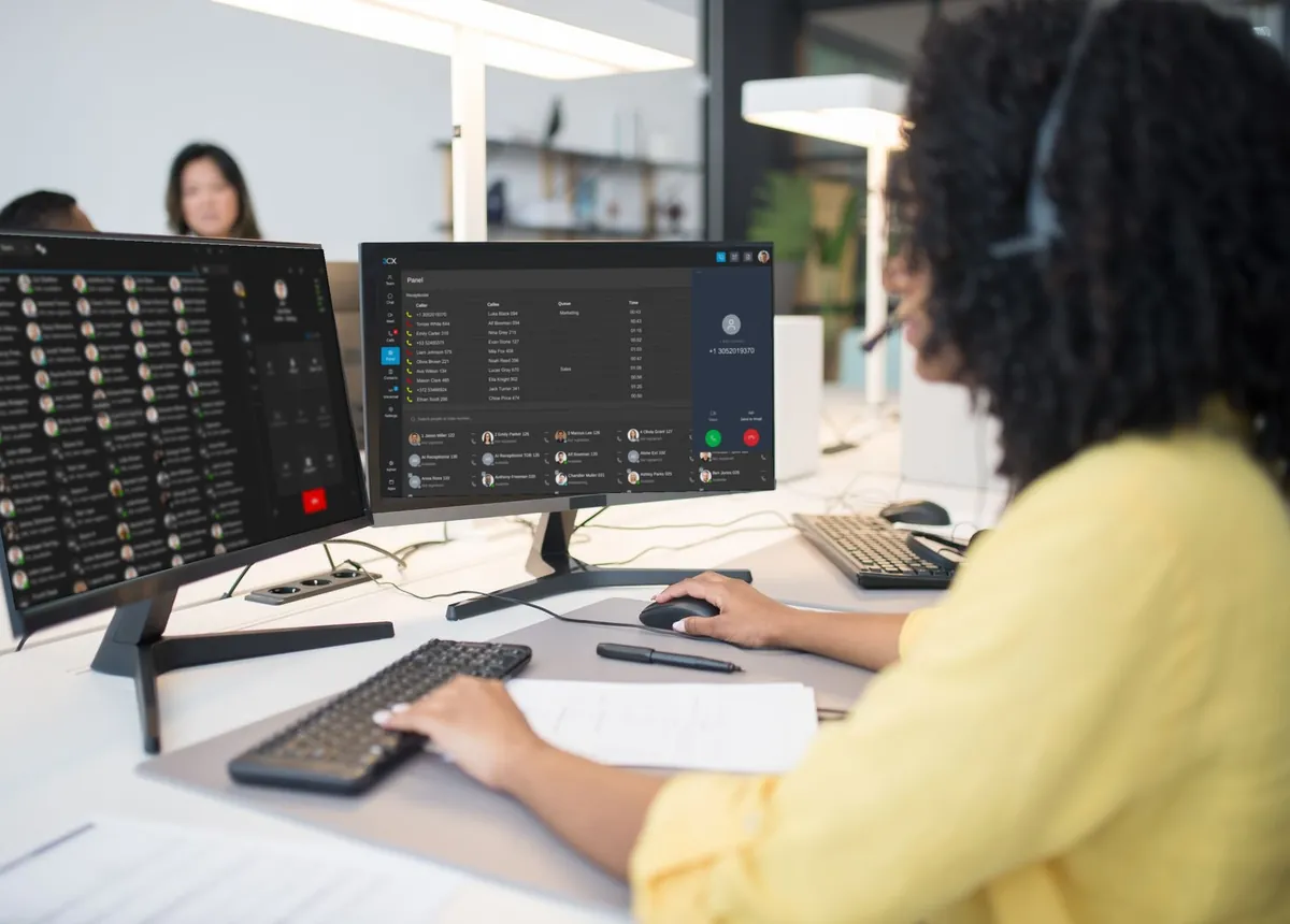 A call-centre agent in a headset at her desk, with the 3CX switchboard showing live calls on one screen and her team's availability on the other.